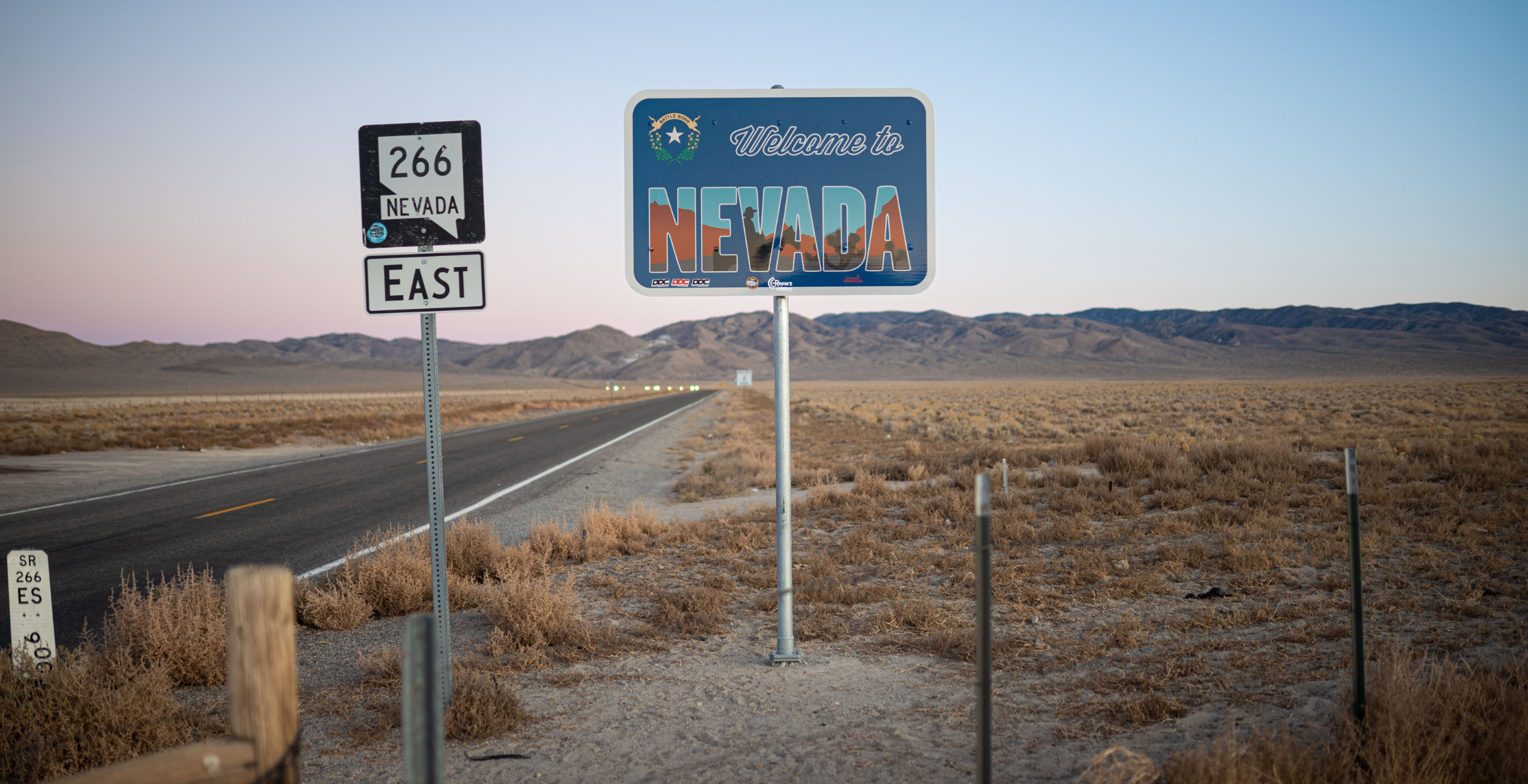 an empty road and mountains are behind a road sign that says 'Welcome to Nevada'